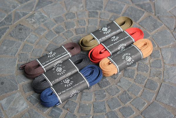 Flat Boot Lace in 54 inch by Alden Madison comes in colors like black, blue, olive, red, and tan. They are neatly arranged on a circular tiled stone surface. Each black and brown lace bundle is wrapped with a sleek black label.