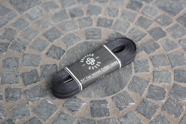 A coiled pair of black and brown laces, wrapped in a branded paper band labeled 54 inch Alden Madison Flat Boot Lace, elegantly rests on a circular stone mosaic surface.