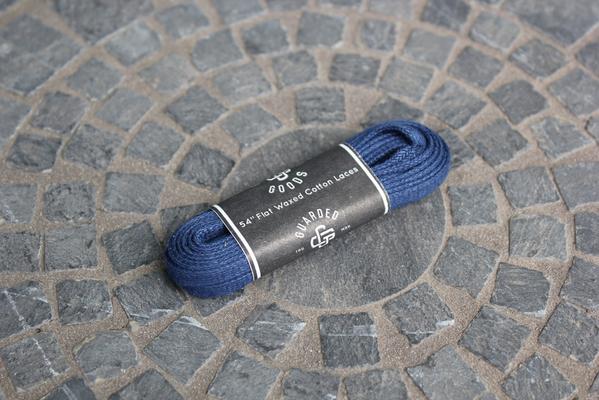 A 54-inch Dark Blue Flat Boot Lace by Alden Madison, wrapped in SAFE and GUARDED labeled black packaging, sits on a textured stone surface with a mosaic pattern reminiscent of Alden shoes detailed craftsmanship.