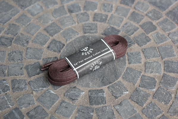 Alden Madisons Flat Boot Lace in 54 inches, featuring brown cotton with black and brown accents, is neatly wrapped in a sleek black paper band displaying text and the logo, placed on a circular stone-tiled surface.