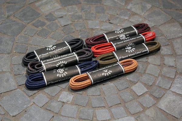 Nine bundles of 27-inch Alden Madison Laces - Shoe Lace Guarded Goods in black, brown, tan, red, olive, and navy are arranged on tiled stone. Each bundle is wrapped with a branded paper label.