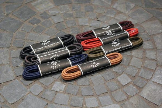 Nine bundles of 27-inch Alden Madison Laces - Shoe Lace Guarded Goods in black, brown, tan, red, olive, and navy are arranged on tiled stone. Each bundle is wrapped with a branded paper label.