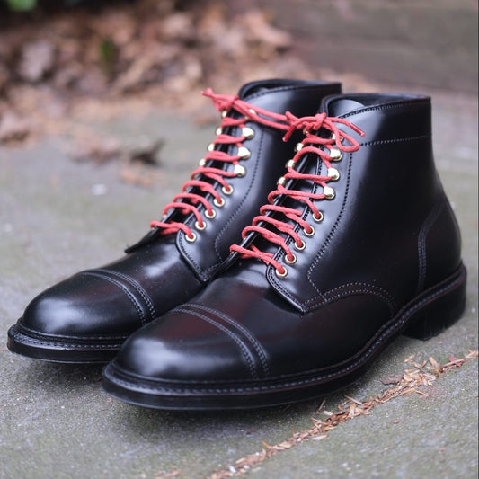A pair of Alden Madisons D9867HC- Stitchup 2/2 Boot in Black Shell Cordovan, featuring polished black leather and red laces with eyelets, rests on a concrete surface. The rugged Commando sole enhances durability, with dry leaves in the background adding a Stitchdown.com style aesthetic.