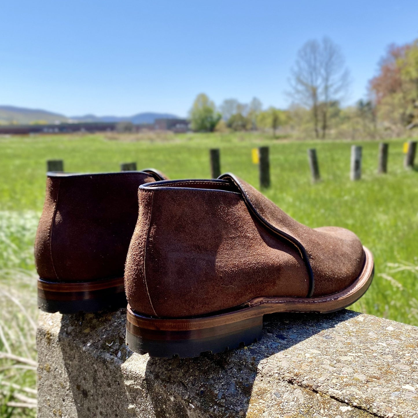 A pair of Alden Madison D8704C - Folsom Slip On Chukka in Tobacco Reverse Chamois with Commando Rubber Soles sits on a stone ledge outdoors, with green grass, trees, and blue sky in the background.