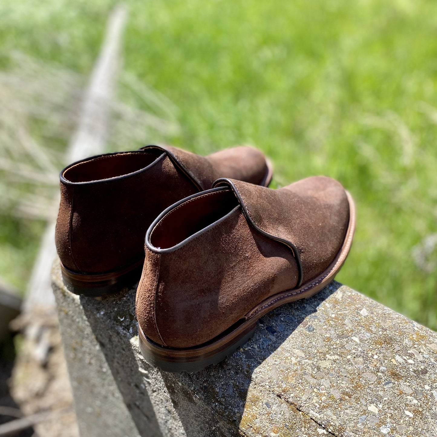 The Alden Madison D8704C Folsom Slip On Chukka in Tobacco Reverse Chamois, featuring rugged commando rubber soles, sits on a concrete ledge outdoors with blurred green grass in the background.