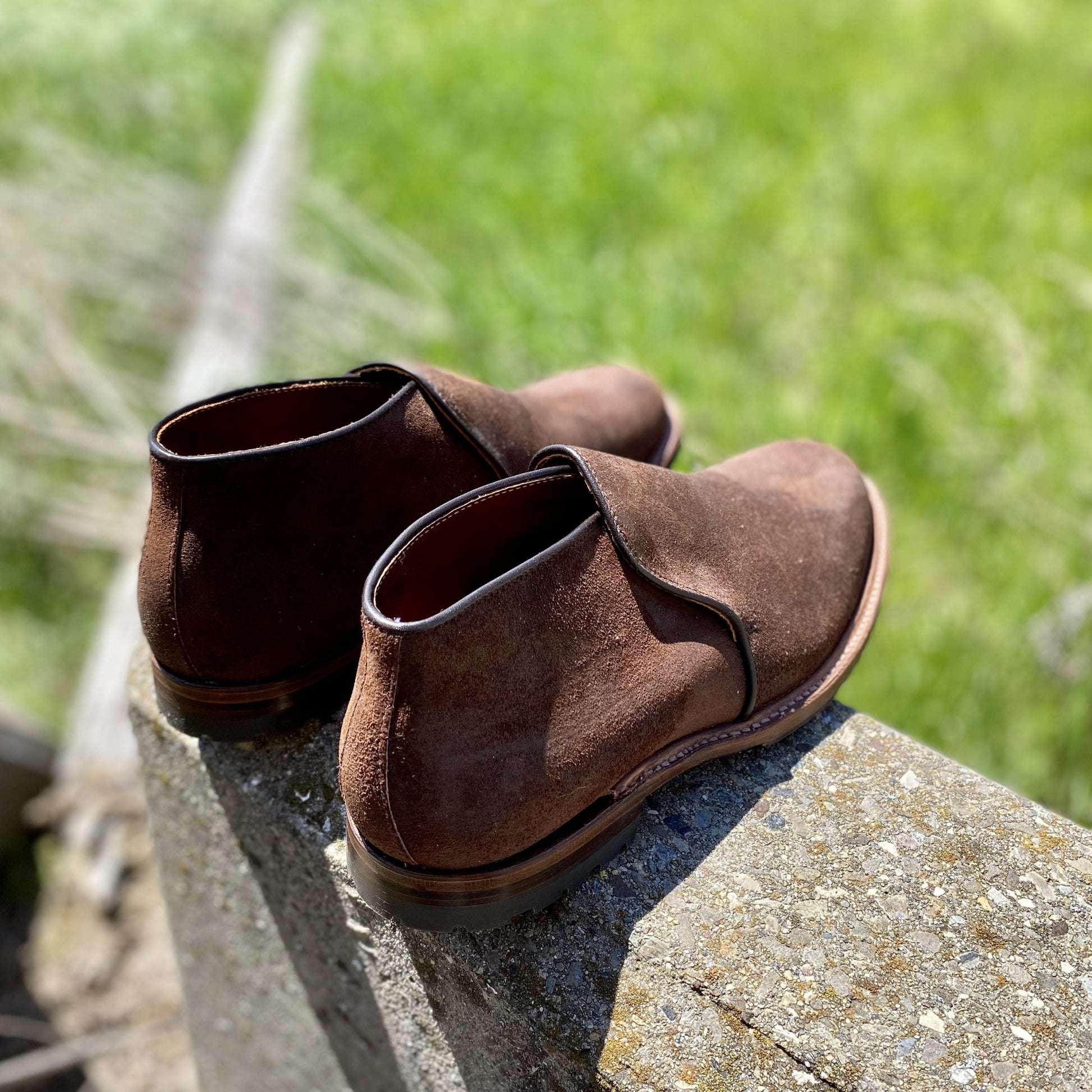 The Alden Madison D8704C Folsom Slip On Chukka in Tobacco Reverse Chamois, featuring rugged commando rubber soles, sits on a concrete ledge outdoors with blurred green grass in the background.