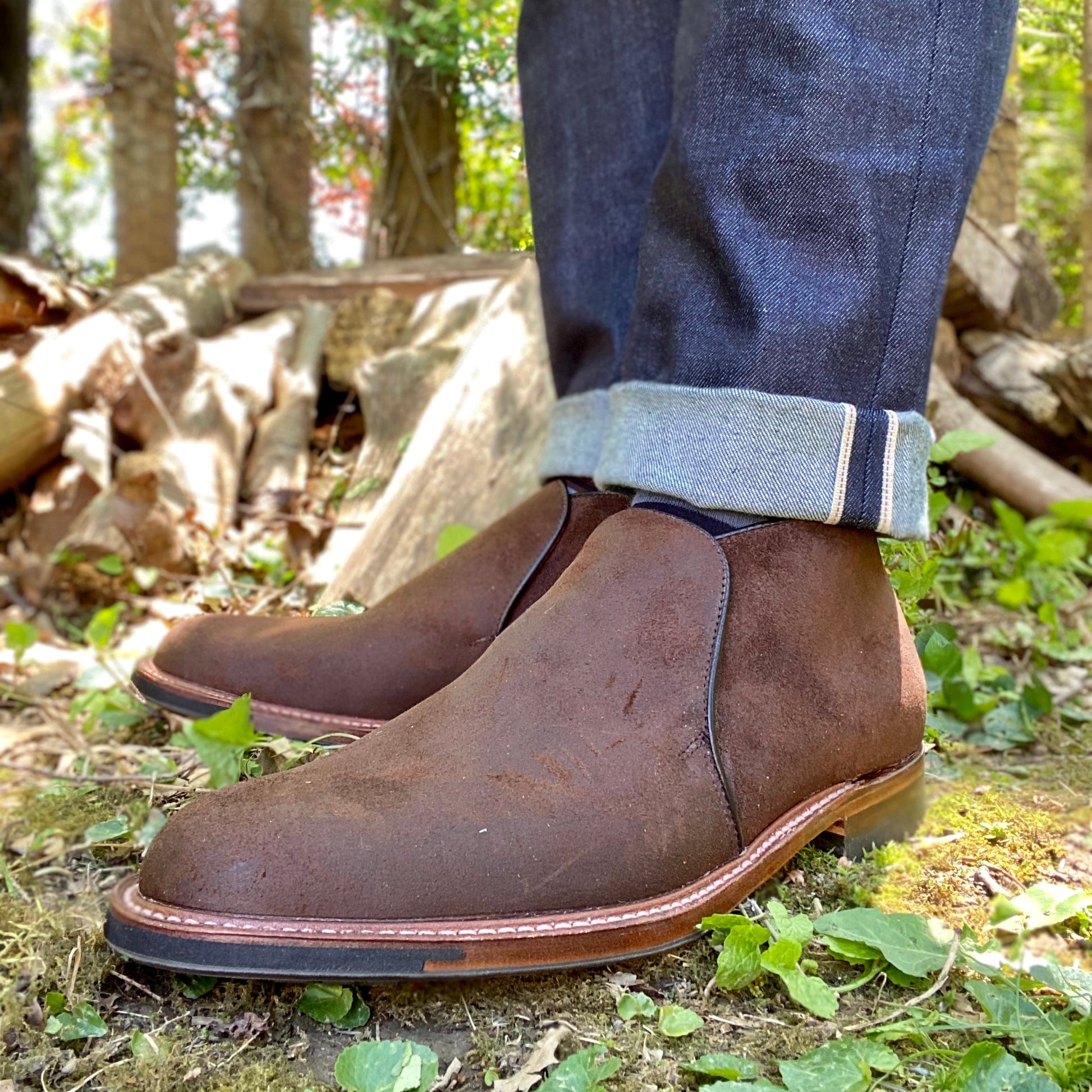 Close-up of a person wearing Alden Madison D8704C - Folsom Slip On Chukka in Tobacco Reverse Chamois with cuffed dark blue jeans, standing on grass and leaves outdoors with logs and trees in the background.