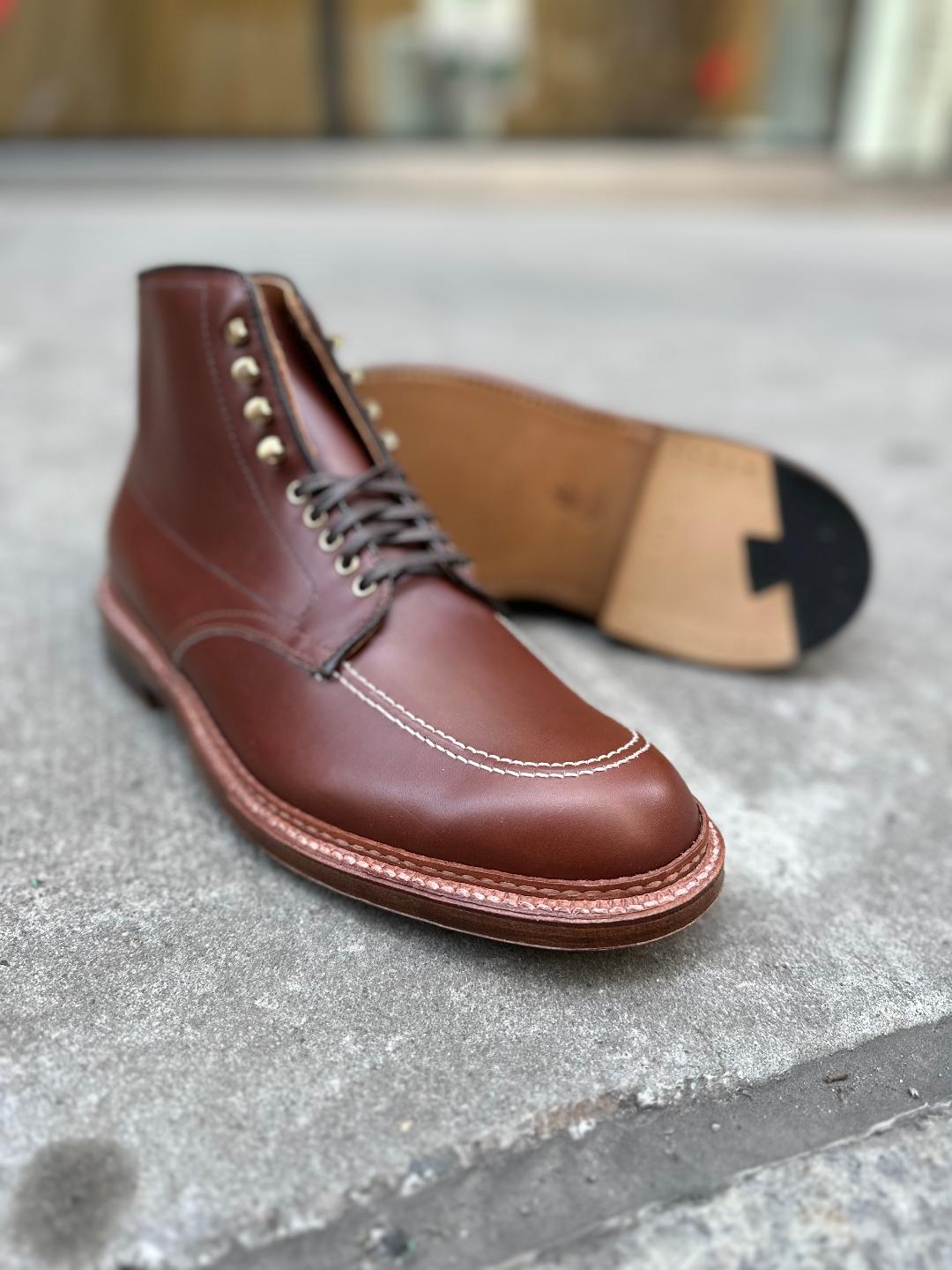 Close-up of an Alden Madison D2940H - Madison Indy Boot in Madison Brown Calf, showcasing white stitching, metallic eyelets, and black laces. The boot rests on a textured gray surface with its sole visible in the background.