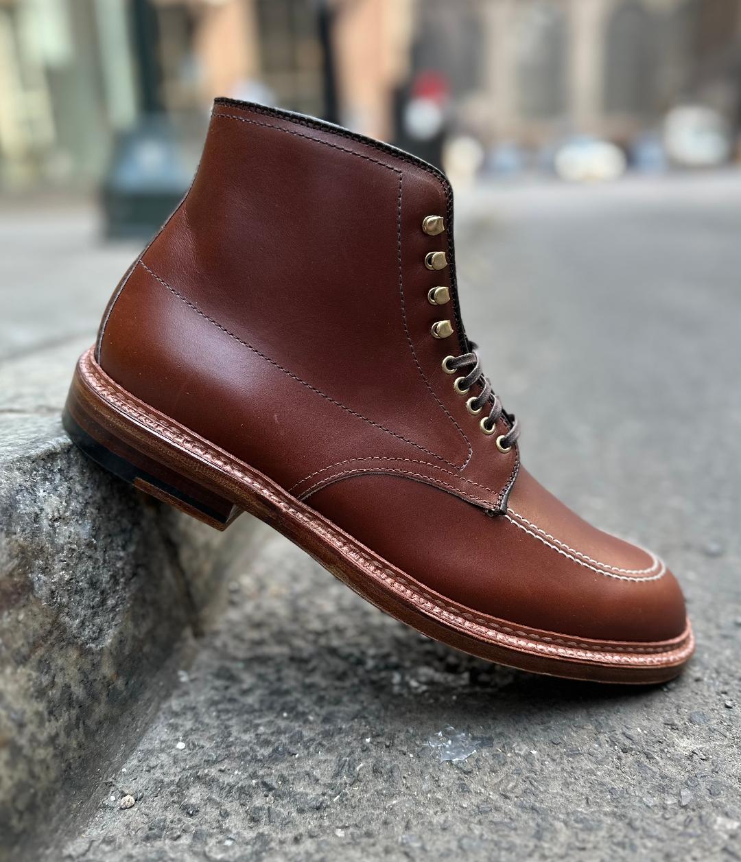 An Alden Madison D2940H - Madison Indy Boot in rich Madison Brown calfskin stands on concrete, showcasing a lace-up design and contrasting stitching against a city street backdrop with blurred buildings and cars for an urban vibe.