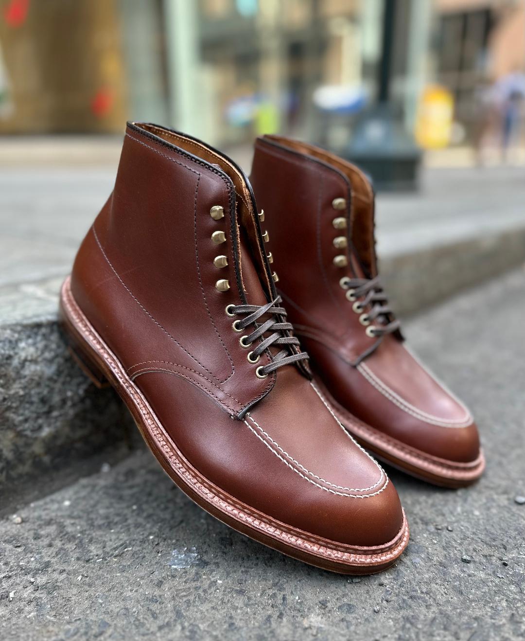 A pair of Alden Madison D2940H - Madison Indy Boots in Madison Brown Calf with brass eyelets and intricate stitching rest on a stone curb, with a slightly blurred urban street setting enhancing their craftsmanship.