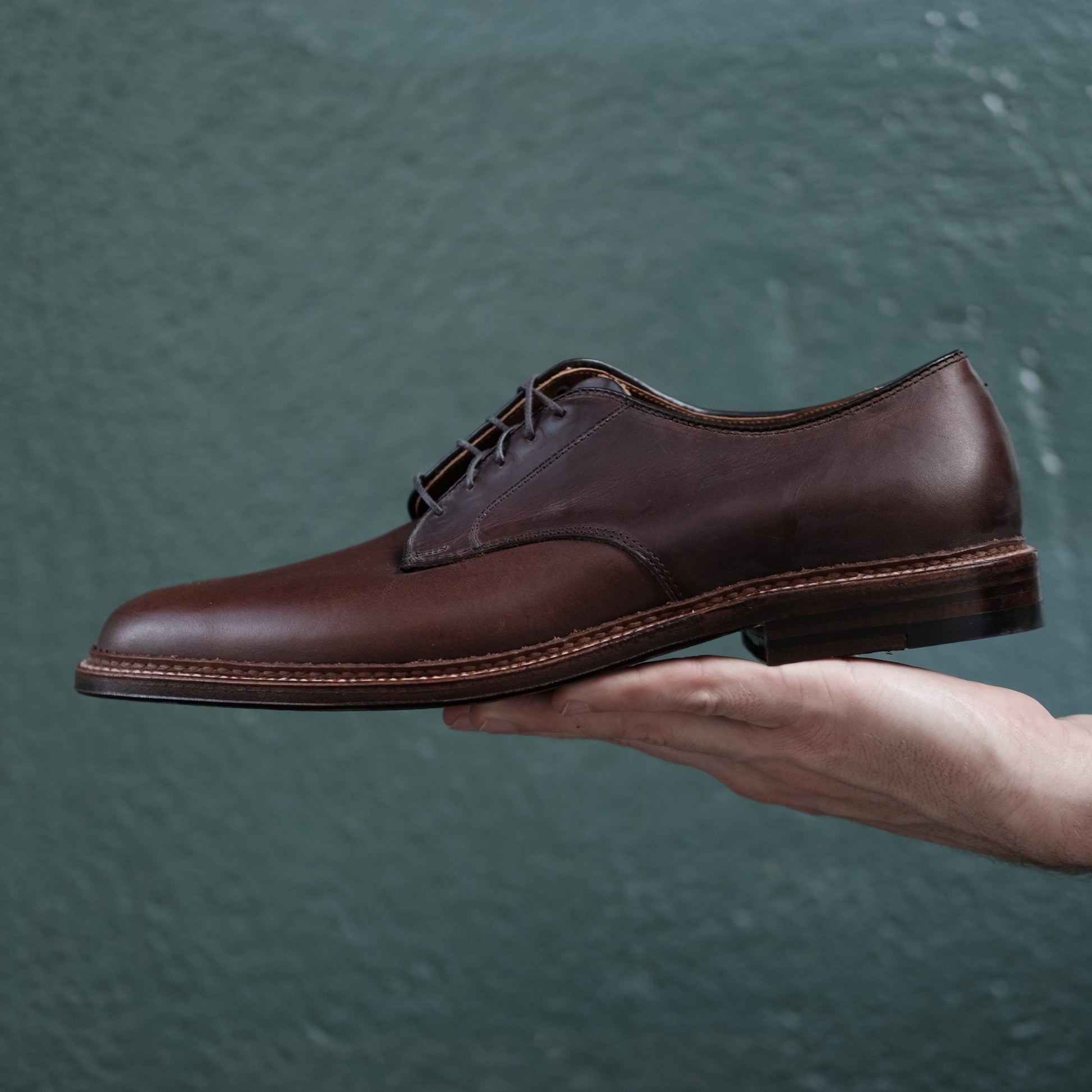 A hand holds up the Alden Madison 29364F - Unlined Plain Toe Dover in Brown Chromexcel, featuring a leather sole, against a green textured background.