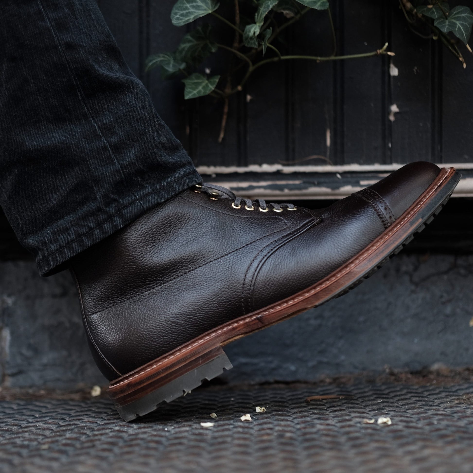 A close-up shows a person in Alden Madisons D9819HC - Jumper Boot in Regina Brown Grain standing on a patterned metal surface. The boots, crafted from premium calfskin, complement sleek dark pants above them. Lush green foliage peeks through in the background.