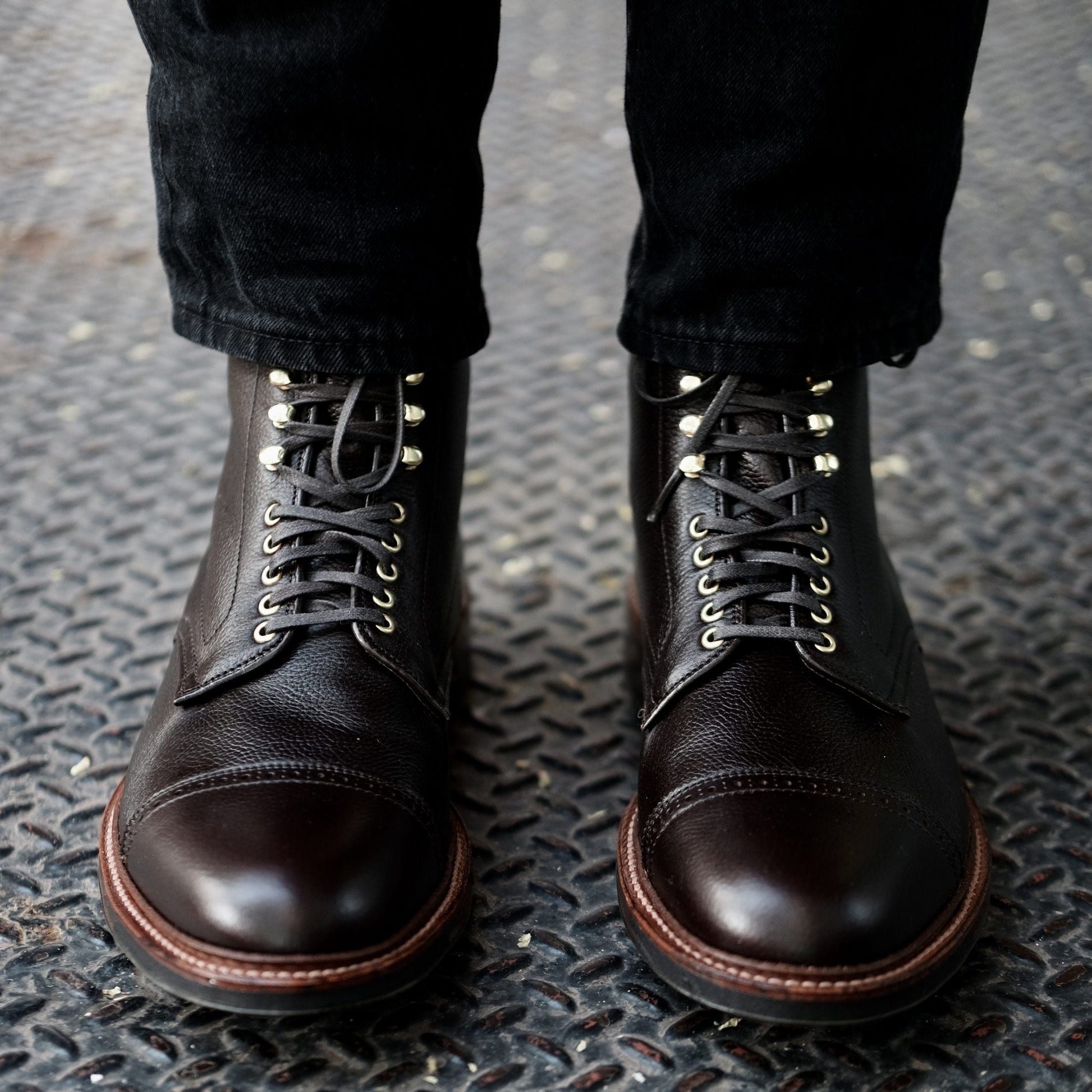 Dark brown Alden Madison D9819HC Jumper Boots in Regina Brown Grain calfskin leather, worn over black pants, feature metal eyelets, a smooth toe cap, and a rugged Commando Sole resting on a textured steel surface.