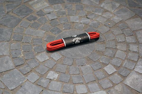 A 27-inch coiled red shoelace from Alden Madison's Laces - Shoe Lace Guarded Goods, with a black and white label, rests on a round, mosaic-patterned stone floor.
