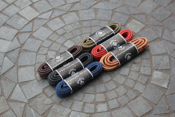 Six pairs of 48-inch Alden Madison Laces - Round Boot Laces in brown, dark green, red, black, navy blue, and tan are neatly coiled on a stone-patterned surface. Each bundle features a branded black paper label.