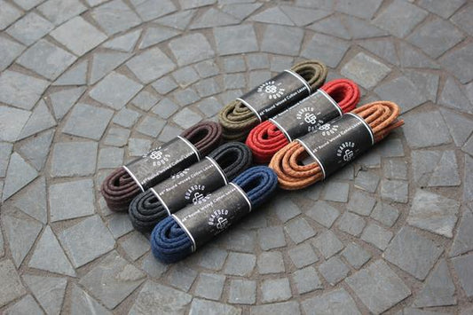 Six pairs of 48-inch Alden Madison Laces - Round Boot Laces in brown, dark green, red, black, navy blue, and tan are neatly coiled on a stone-patterned surface. Each bundle features a branded black paper label.