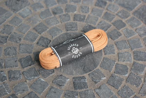 A pair of neatly coiled 54-inch Flat Boot Laces in tan, packaged with Alden Madisons branding, rests on a circular stone-tile surface, perfectly complementing Aldens style.